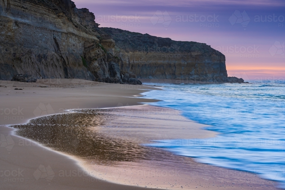 Wave patterns left on wet sand below rugged sea cliffs at twilight - Australian Stock Image