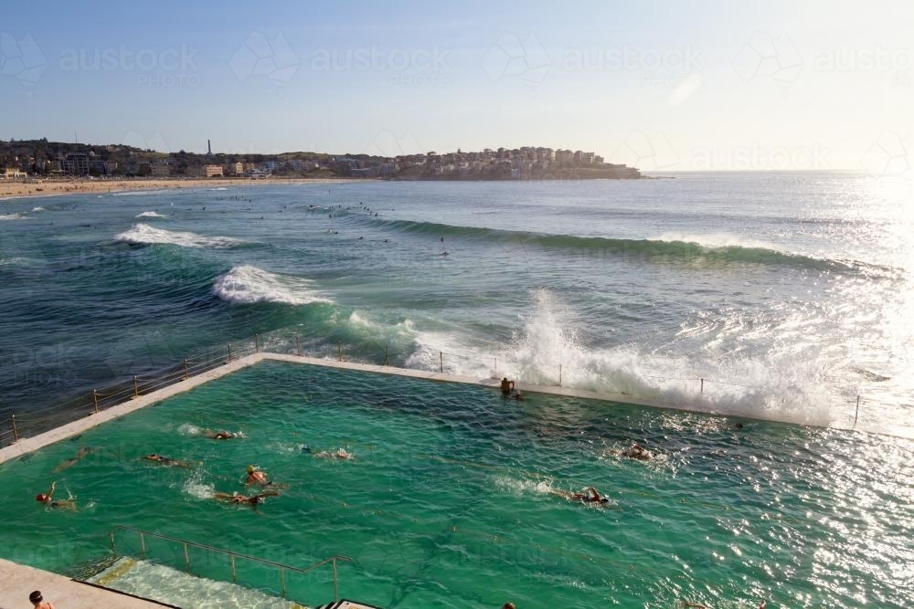 Waves crashing over Bondi Baths at sunrise with people swimming laps - Australian Stock Image