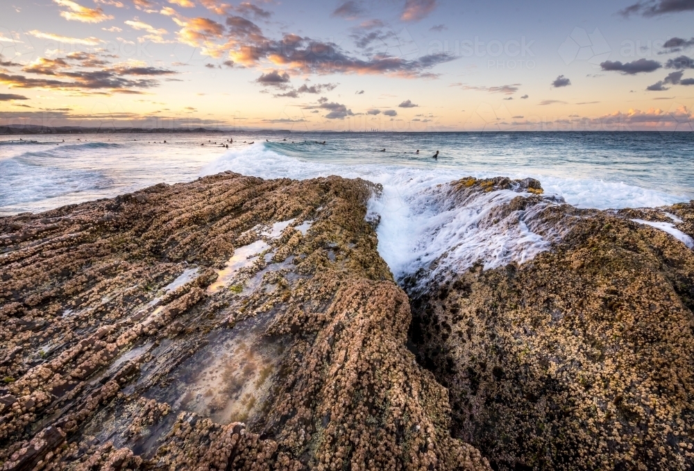 Waves crashing over rocks with surfers in the background at sunset - Australian Stock Image