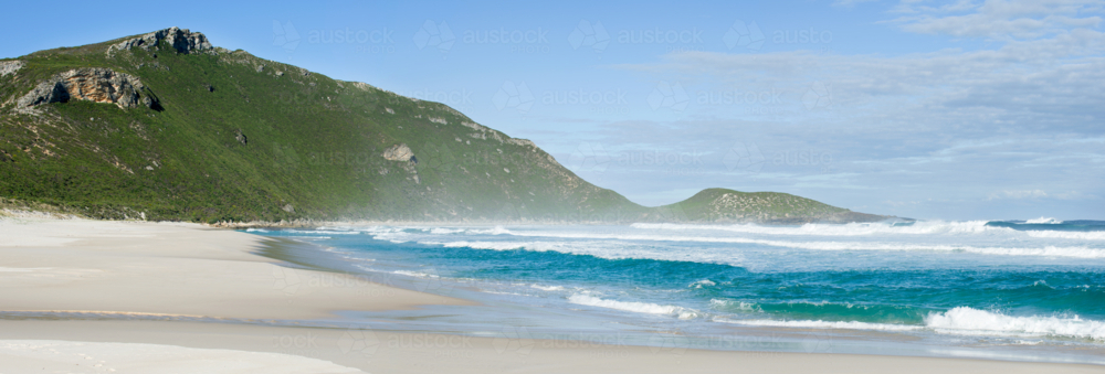Waves roll onto a deserted beach beneath a green headland - Australian Stock Image
