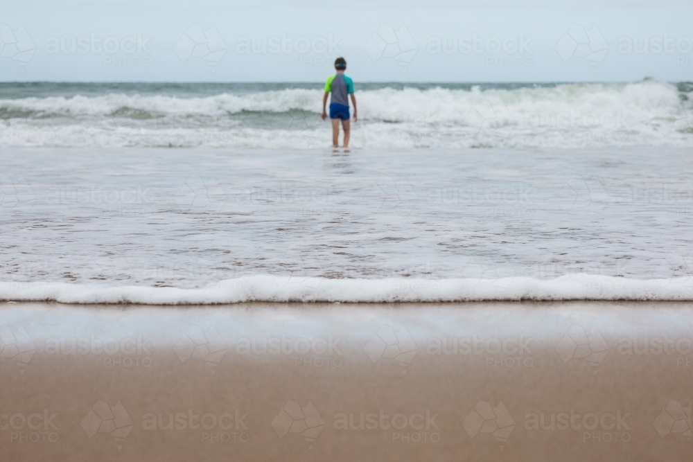Waves rolling in on sand at beach with young boy walking out to ocean - Australian Stock Image