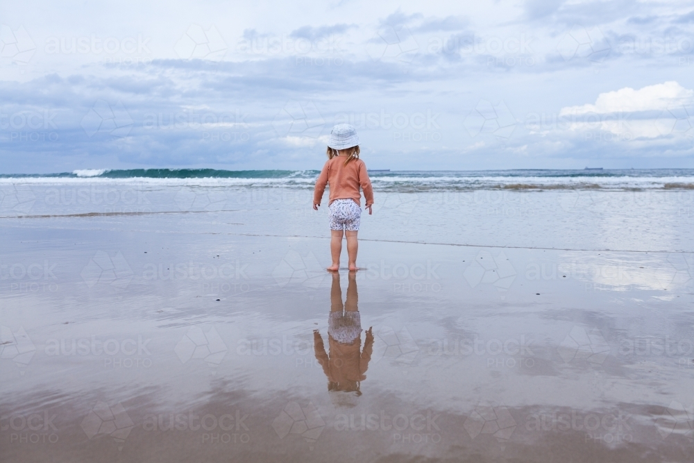 Wet sand reflecting little Aussie kid looking towards waves at beach on winters day - Australian Stock Image