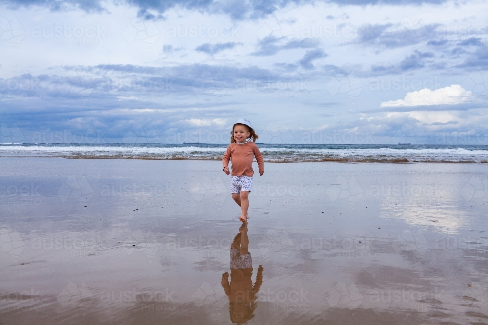 Wet sand reflecting little Aussie kid running away from waves at beach on winters day - Australian Stock Image