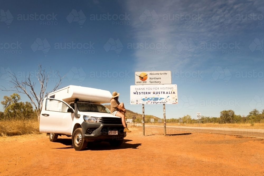 White camper van and a woman sitting on the hood looking at the signage beside her in a rural area - Australian Stock Image