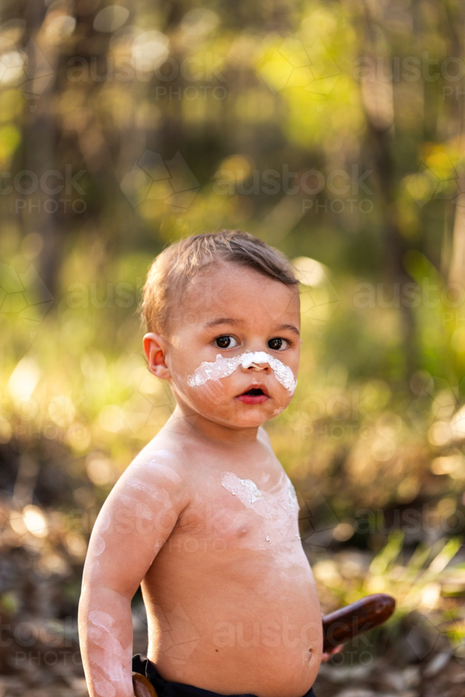 White ochre paint on Aboriginal child’s face in natural bushland with backlighting - Australian Stock Image