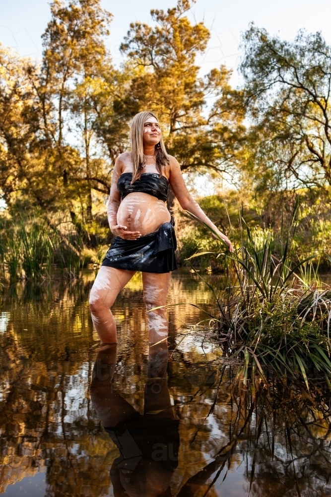 Wide shot of Aboriginal Australian woman standing in creek water in traditional body paint - Australian Stock Image