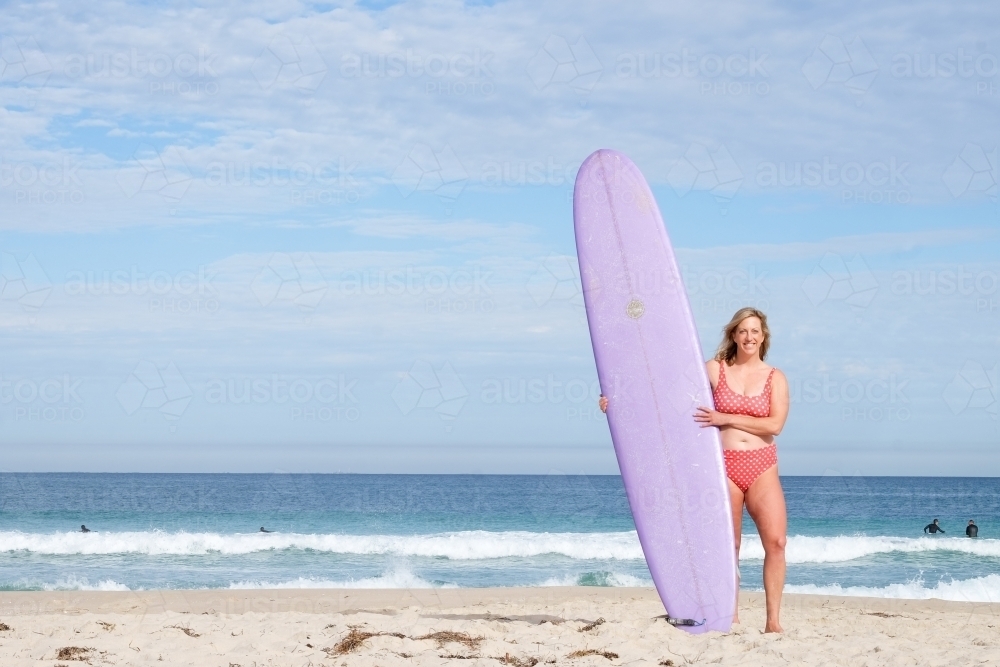 Wide view of woman surfer standing on beach in bikini holding a longboard surfboard - Australian Stock Image