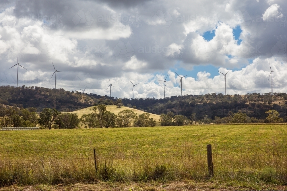 Wind turbines on top of hill in distance on farm with cloudy sky - Australian Stock Image