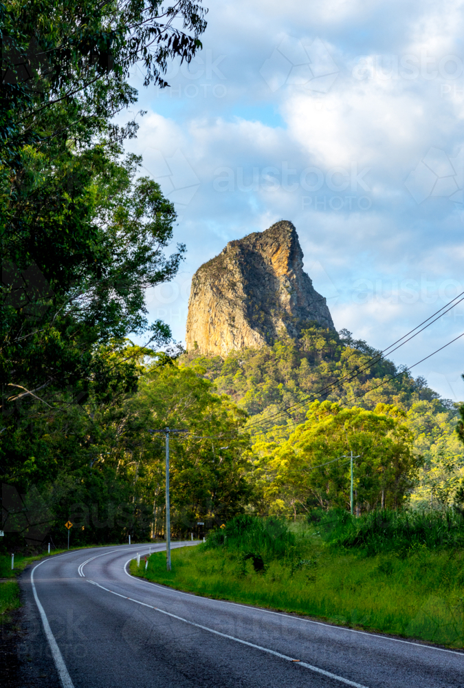 Winding bitumen road with Mount Tibrogargan lit by sun in background - Australian Stock Image