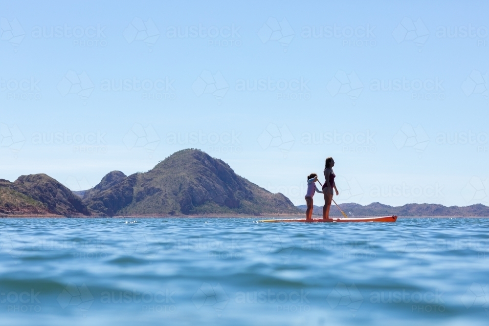 woman and child standing on paddle board in blue water with hills behind - Australian Stock Image