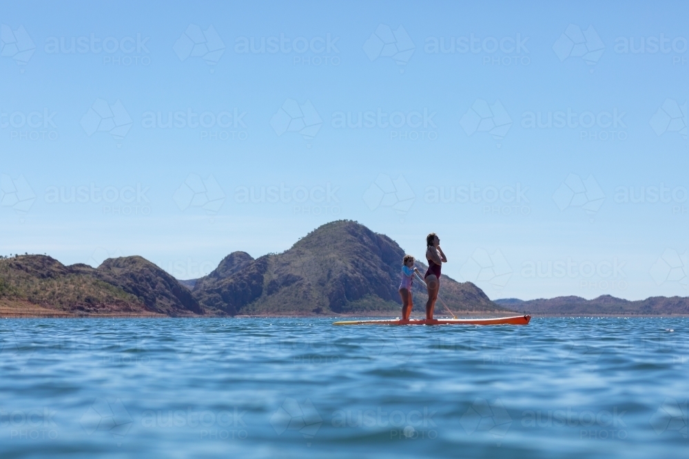 woman and child standing on paddle board in blue water with hills behind - Australian Stock Image