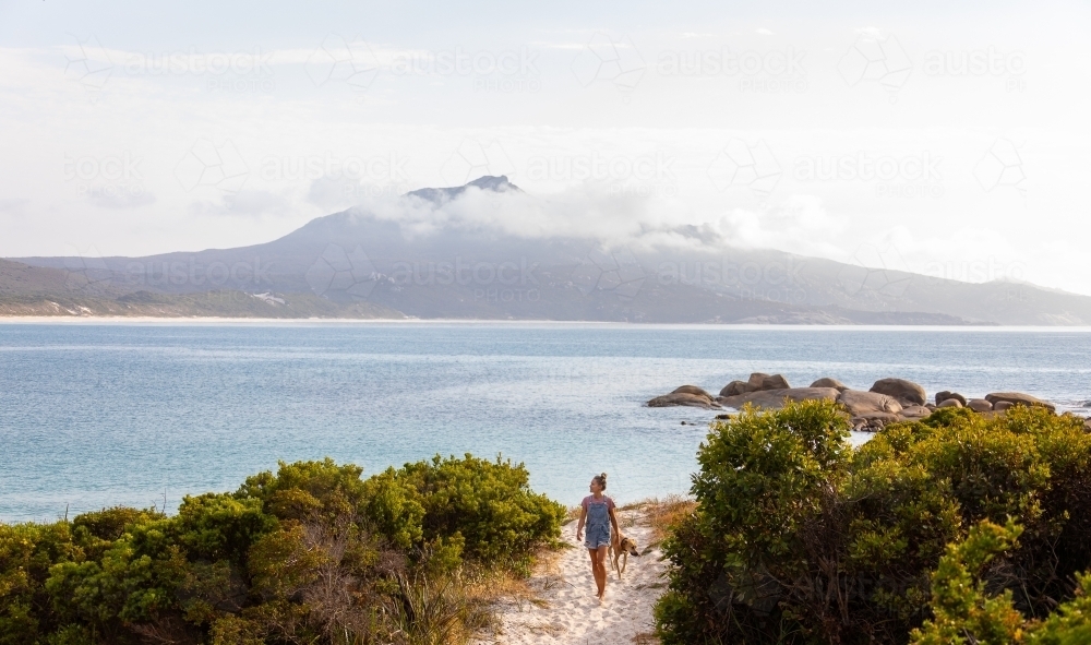 woman and dog walking on beach access path with could-covered Mount Manypeaks in background - Australian Stock Image