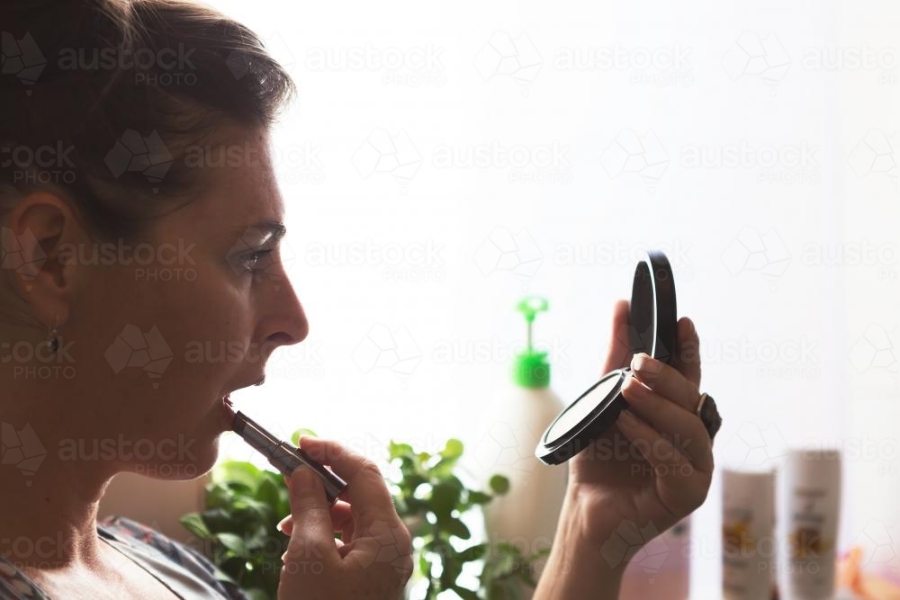 Woman applying lipstick in bathroom with backlit window - Australian Stock Image