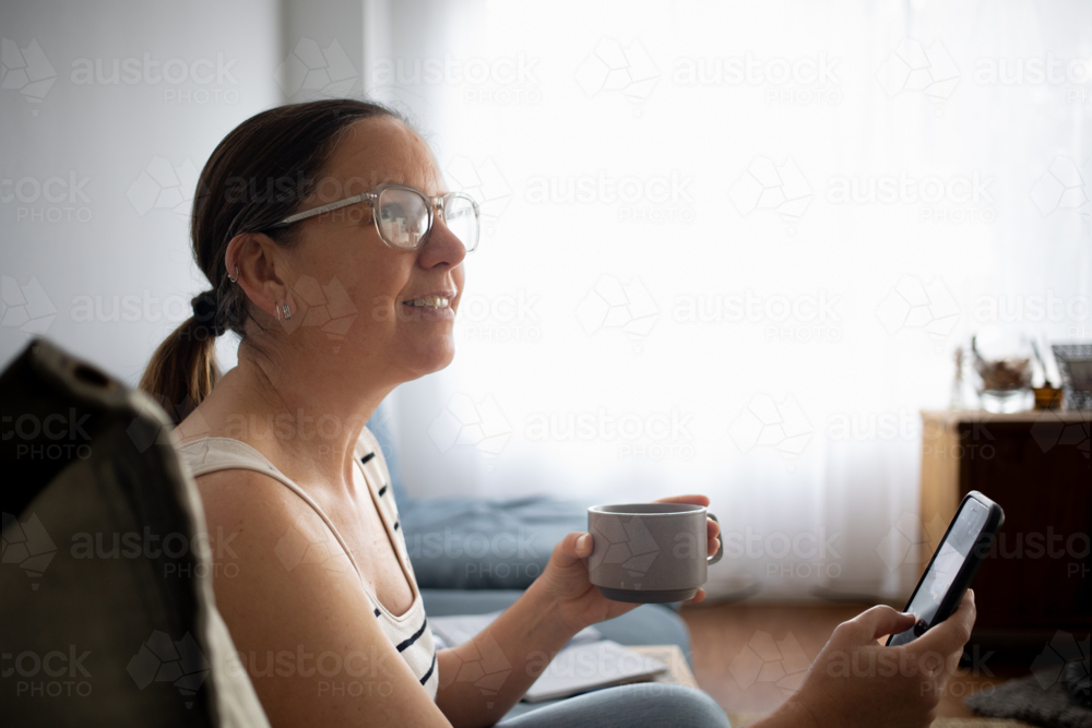 Woman at home looking at someone while using smart phone and drinking coffee - Australian Stock Image