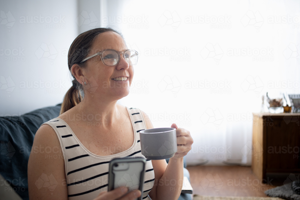 Woman at home on couch smiling at someone while using smart phone and drinking coffee - Australian Stock Image
