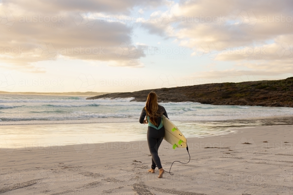 woman carrying surfboard out into the surf from white sand beach - Australian Stock Image