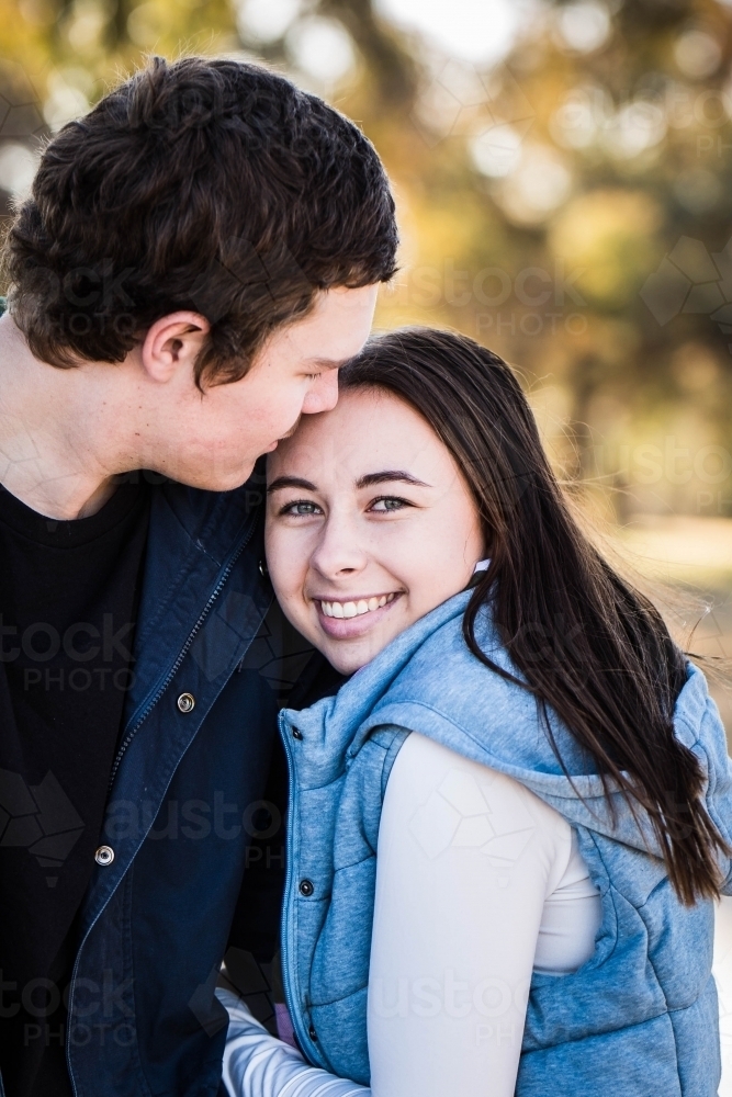 Woman cuddling boyfriend's arm smiling while he kisses her forehead - Australian Stock Image