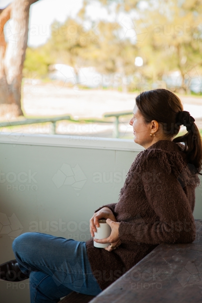 Woman drinking a hot drink on a picnic bench at a park - Australian Stock Image