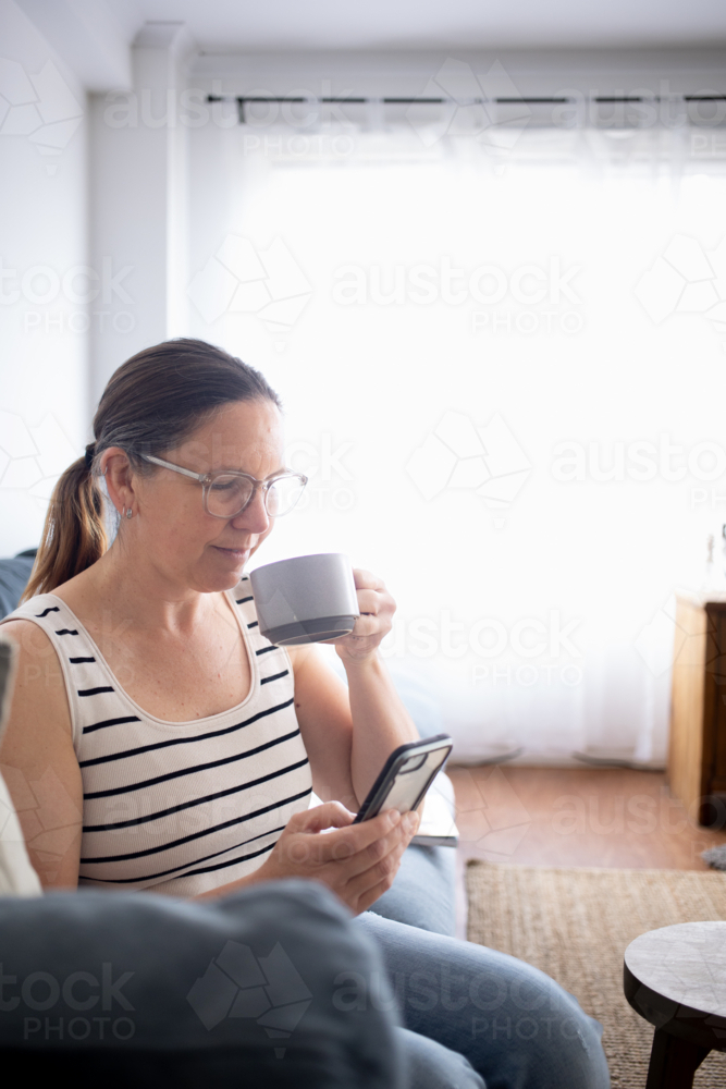 Woman drinking coffee on couch and using smart phone - Australian Stock Image