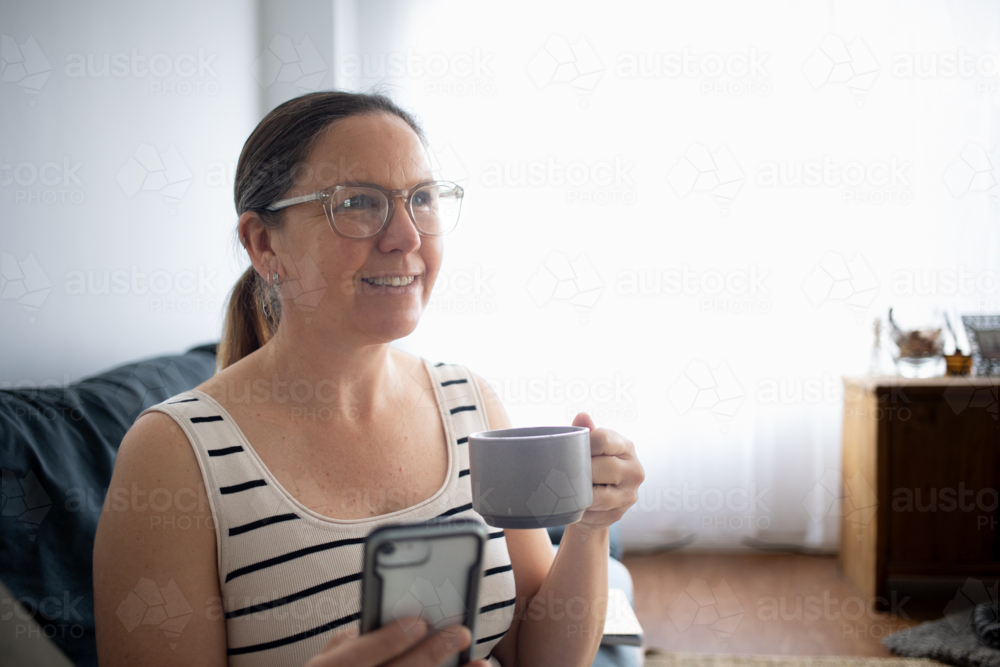 Woman drinking coffee on couch looking off to the side - Australian Stock Image