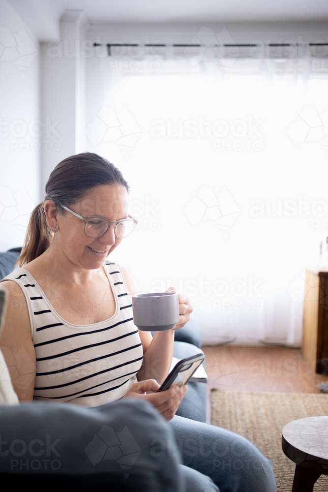 Woman drinking coffee on couch using smart phone - Australian Stock Image