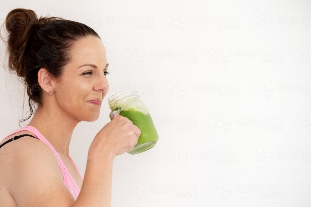 woman drinking green juice smiling in workout gear white background - Australian Stock Image