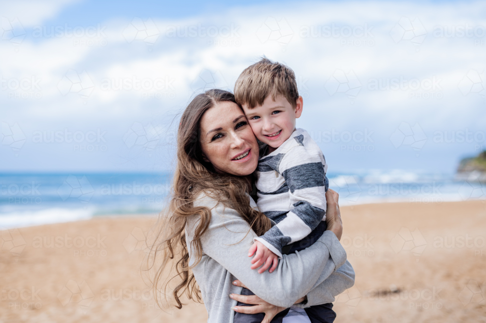 Woman embraces her smiling son at the beach with waves and sand in the background - Australian Stock Image