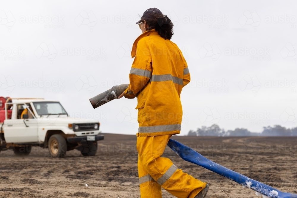 Woman firefighter walking along burnt paddock holding hose - Australian Stock Image