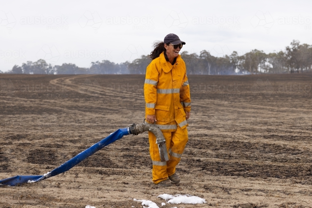 Woman firefighter walking with hose laughing in burnt paddock - Australian Stock Image