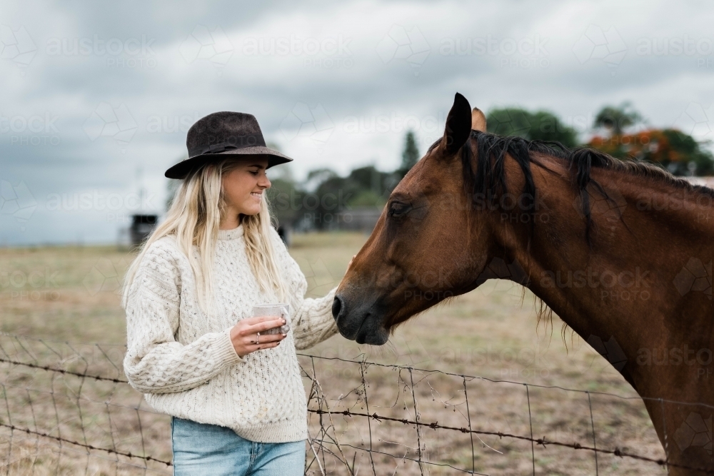 Woman holding a cup of coffee while the pats a horse - Australian Stock Image