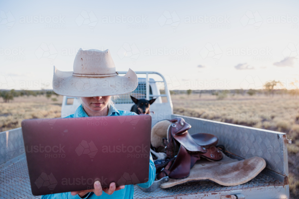 Woman holding a laptop computer sitting at the back of the ute on farm - Australian Stock Image