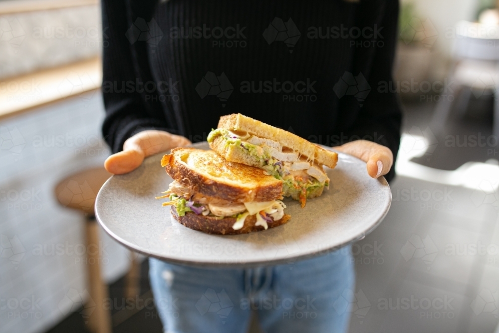 Woman holding a plate with a sandwich cut into half with chicken, avocado and cheese in it - Australian Stock Image