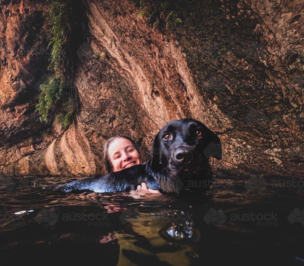 Woman holding black Labrador dog on surface of water hole with gorges wall in background - Australian Stock Image