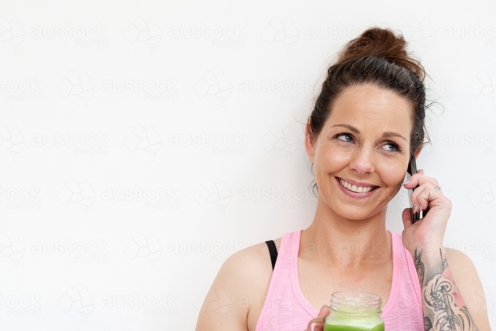 woman holding green juice talking on phone in workout clothes white background - Australian Stock Image