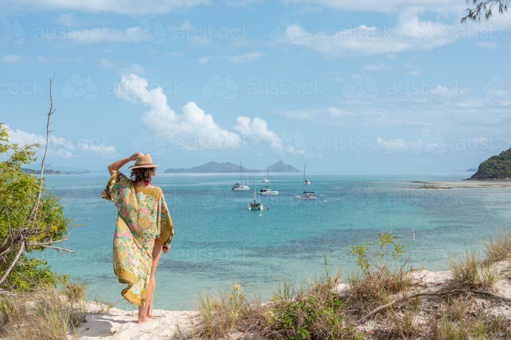 woman holding hat, wearing kimono looking out over calm bay of ocean with islands in background - Australian Stock Image