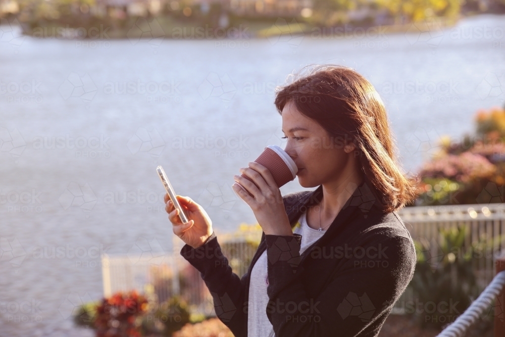 Woman holding mobile and drinking by the lake - Australian Stock Image