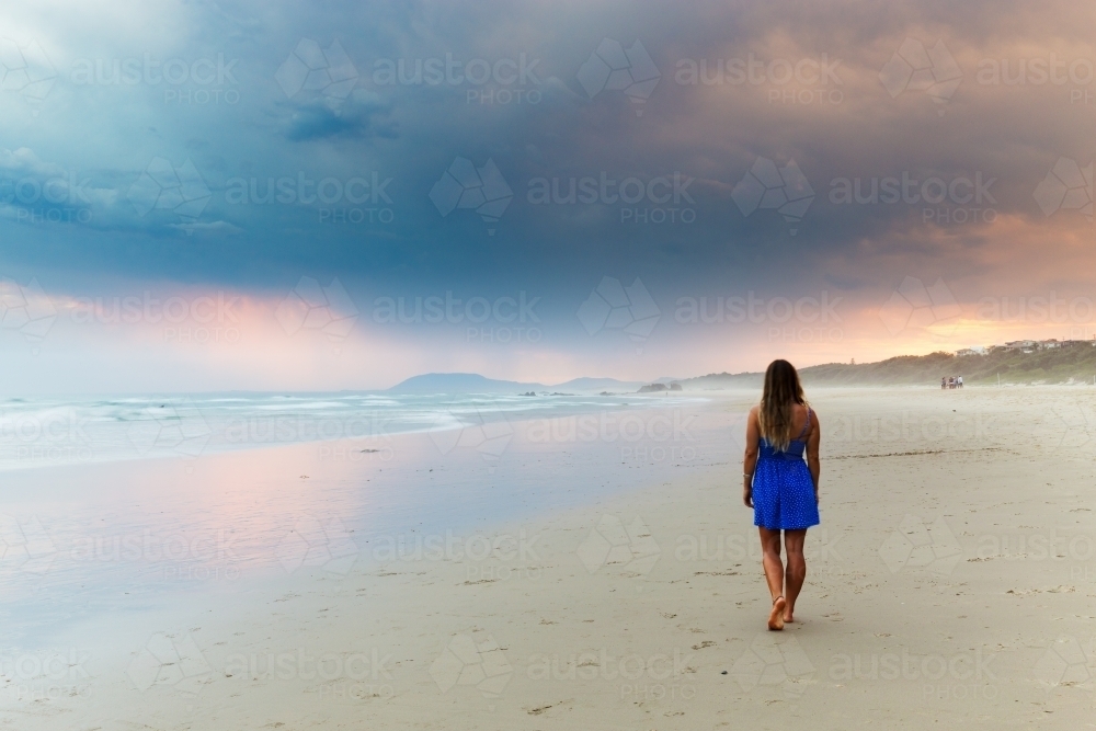 Woman in a blue dress walking on the beach towards a stormy sunset - Australian Stock Image