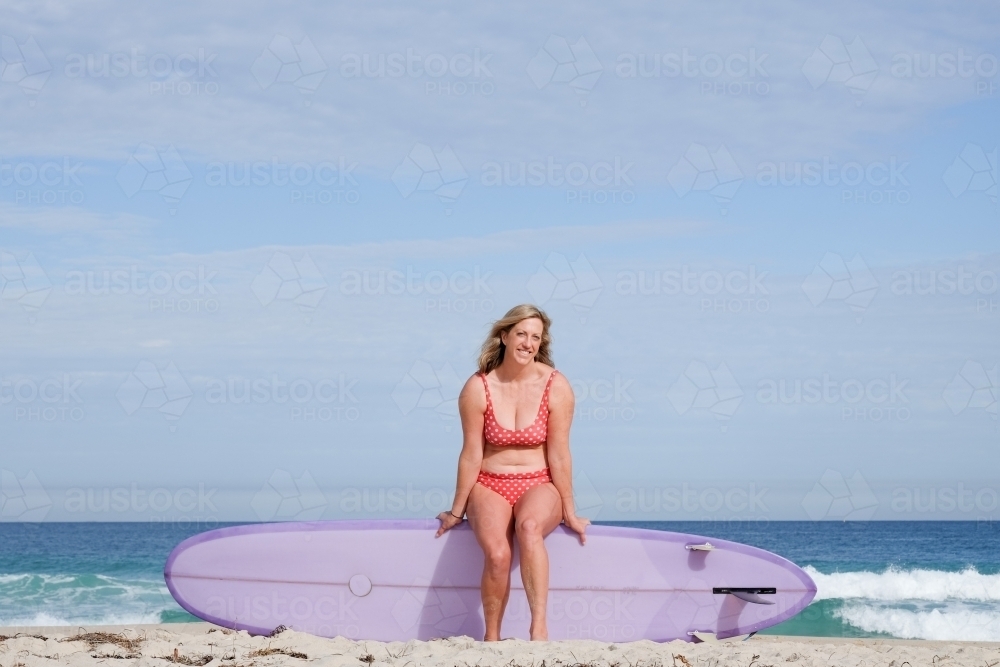 Woman in bikini sitting on longboard surfboard on shoreline of beach looking at camera - Australian Stock Image