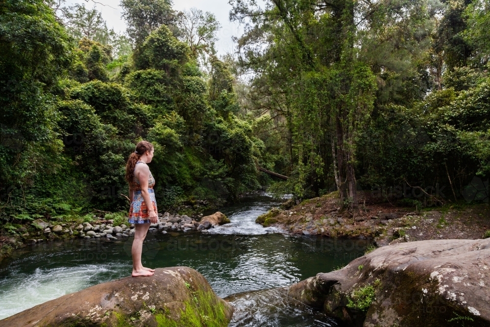 Woman in bright swimmers standing on rock looking out over pool of water - Allyn River - Australian Stock Image