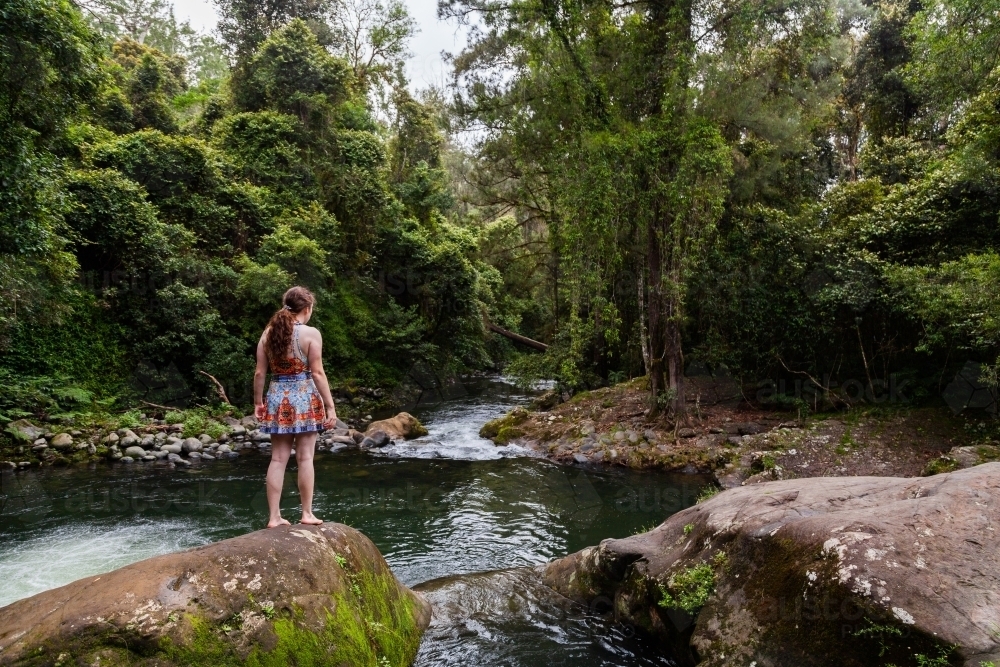 Woman in bright swimmers standing on rock looking out over pool of water - Allyn River - Australian Stock Image