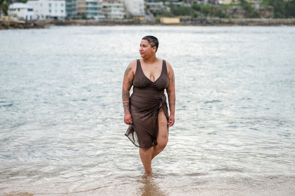 Woman in brown swimsuit walking in shallow waters at the beach - Australian Stock Image