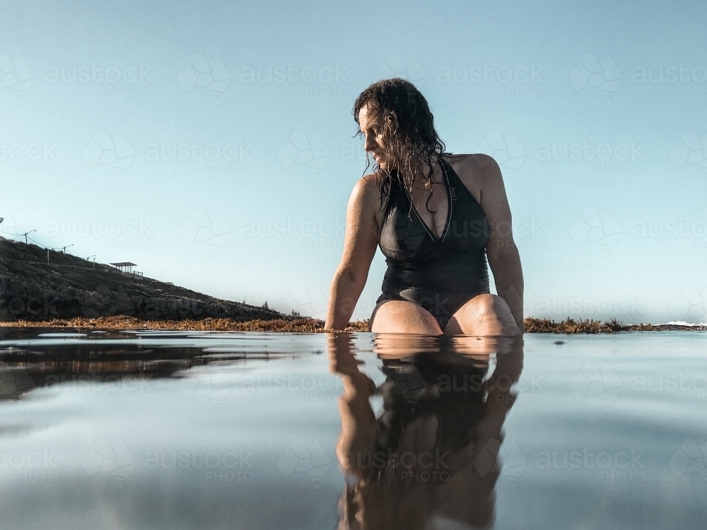 Woman in full piece bathing suit sitting on reef ledge looking into glassy ocean water - Australian Stock Image