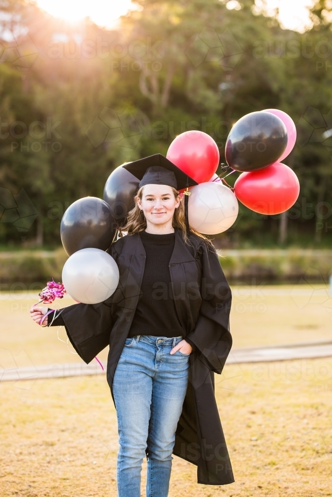 Woman in graduation cap and gown holding balloons standing in park - Australian Stock Image