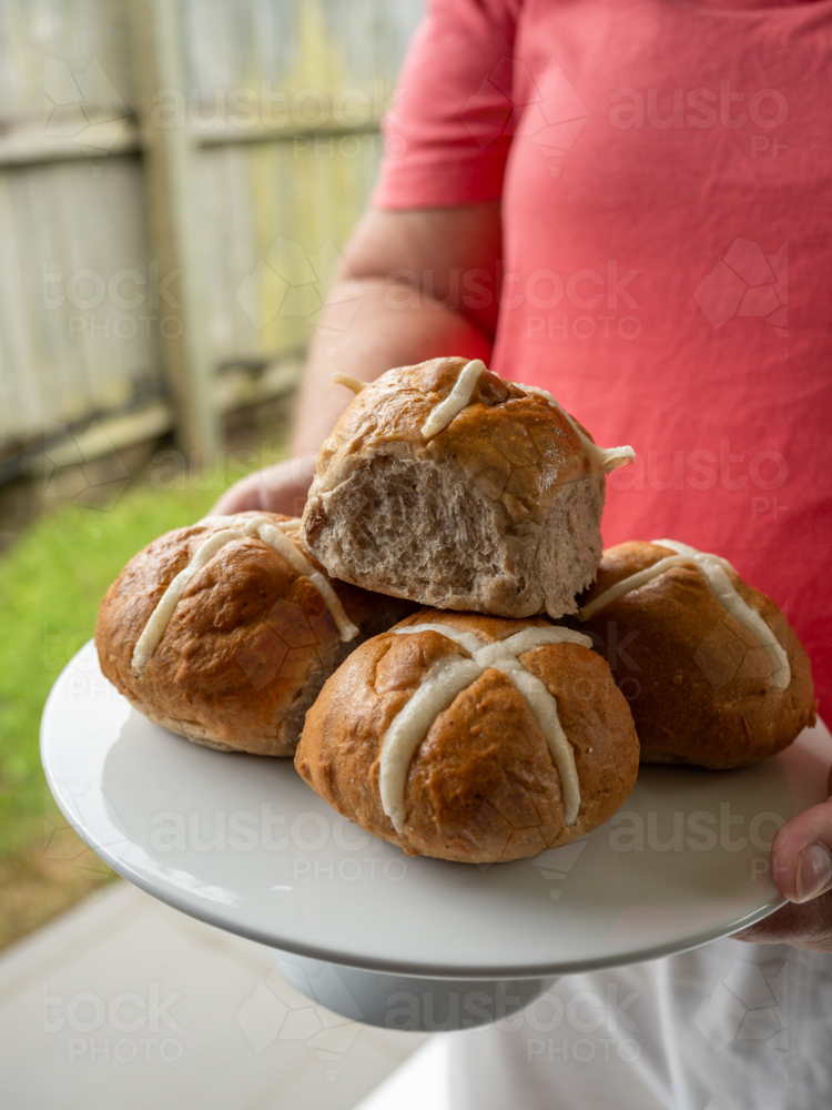 Woman in red shirt holding a cake stand piled with hot cross buns with green grass and wooden fence - Australian Stock Image