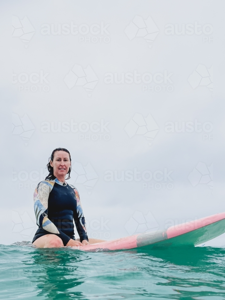 Woman in springsuit sitting on surfboard looking directly on calm overcast day - Australian Stock Image