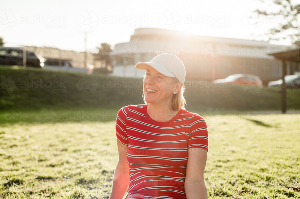 Woman in stripe shirt, sitting in a grassy park under the warm afternoon sun, smiling and relaxed. - Australian Stock Image