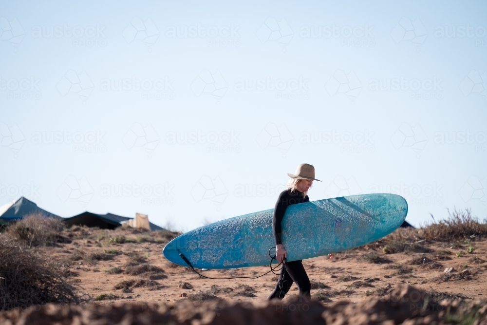 Woman in wetsuit and wide brim hat carrying longboard surfboard through desert like environment - Australian Stock Image