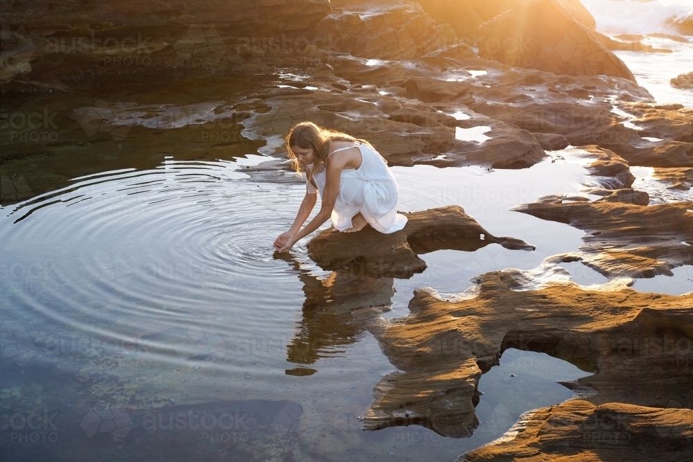 Woman kneeling beside rockpool with golden light and ripples - Australian Stock Image