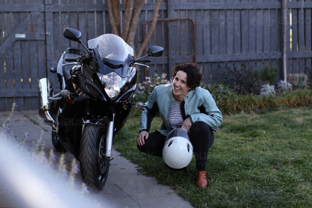 Woman laughing and crouching next to motorbike in garden - Australian Stock Image