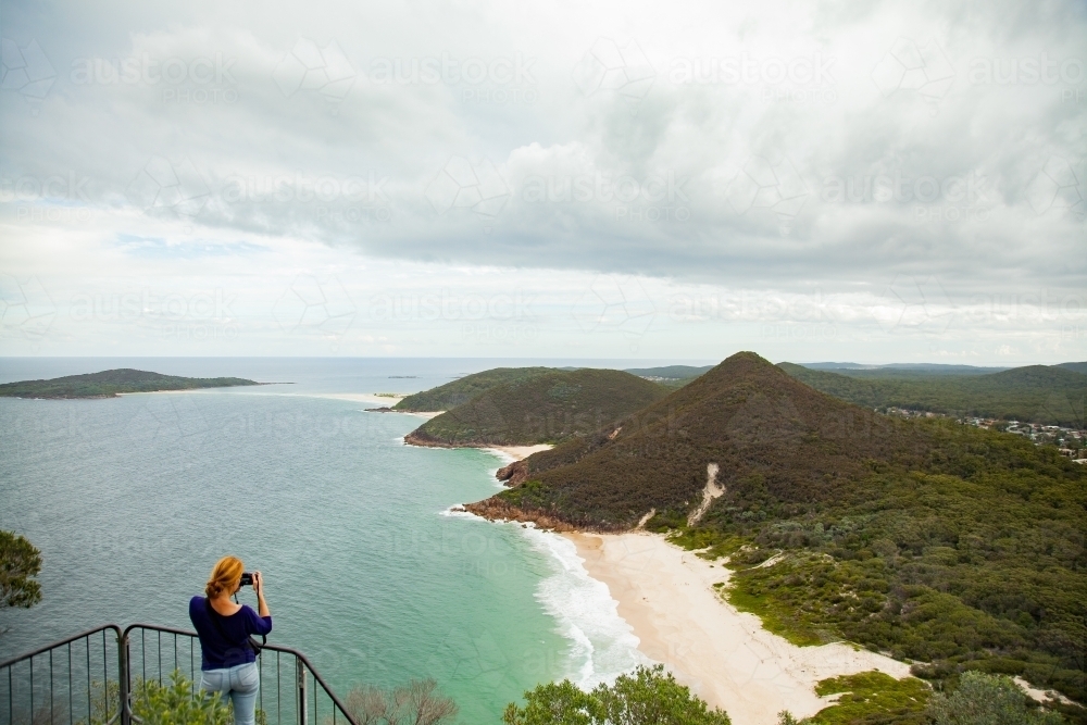 Woman looking out from Tomaree Head Summit walk taking photos of ocean and islands - Australian Stock Image
